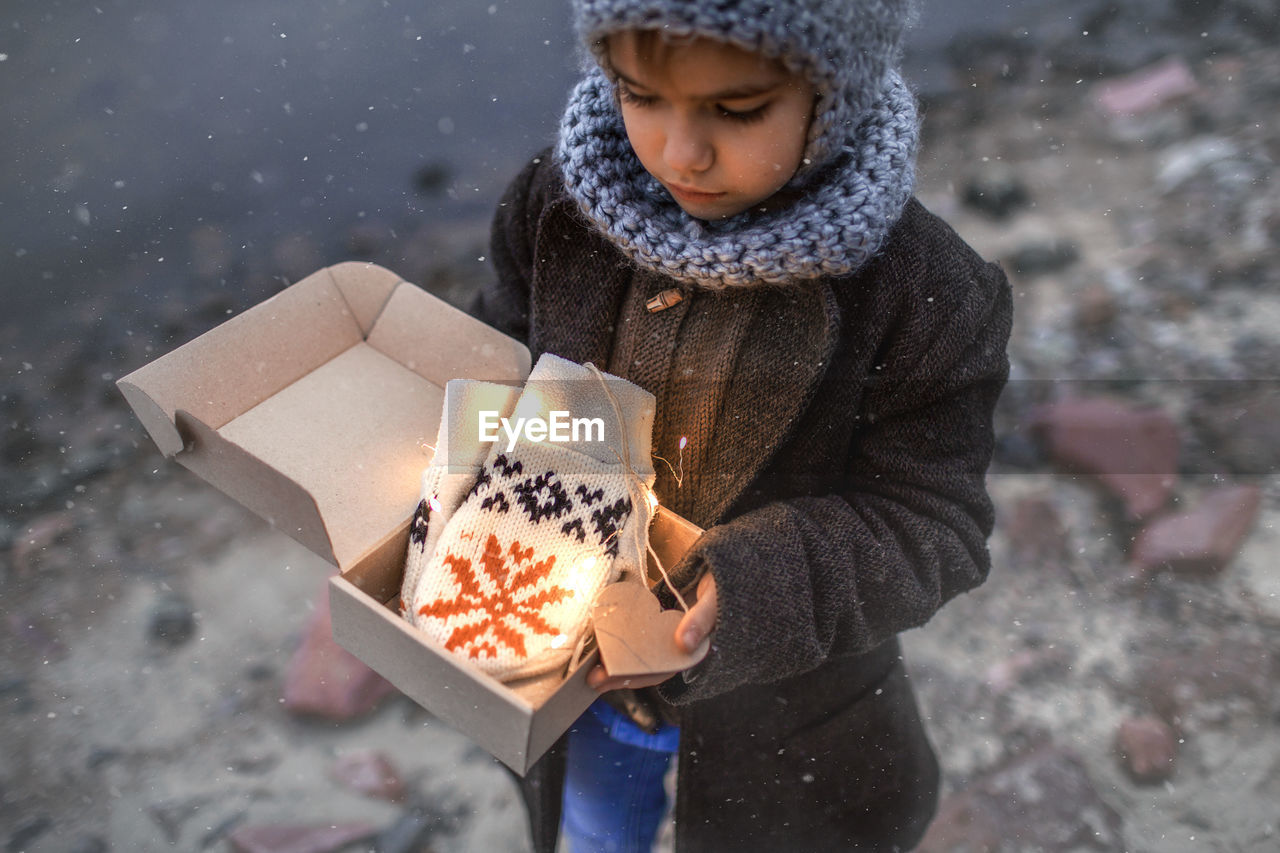 Pretty girl in knitted grey hat opening a crafted gift box with a new pair of gloves, snowfall