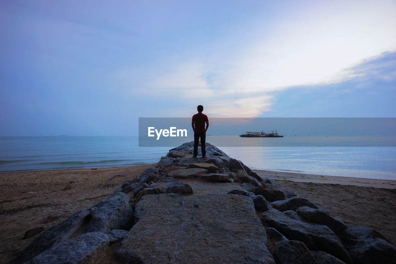 Rear view of man standing on rock at beach against cloudy sky during sunset