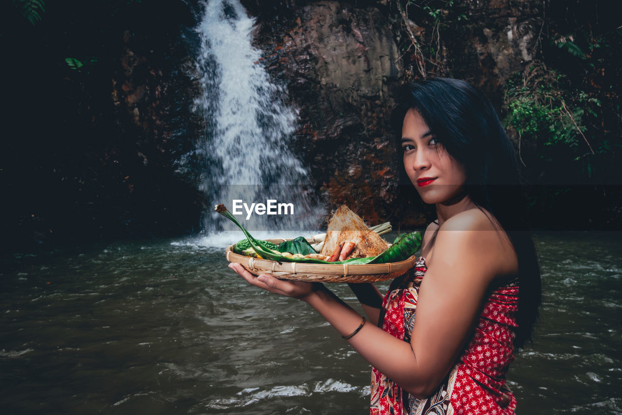 Portrait of woman holding food in tray against waterfall