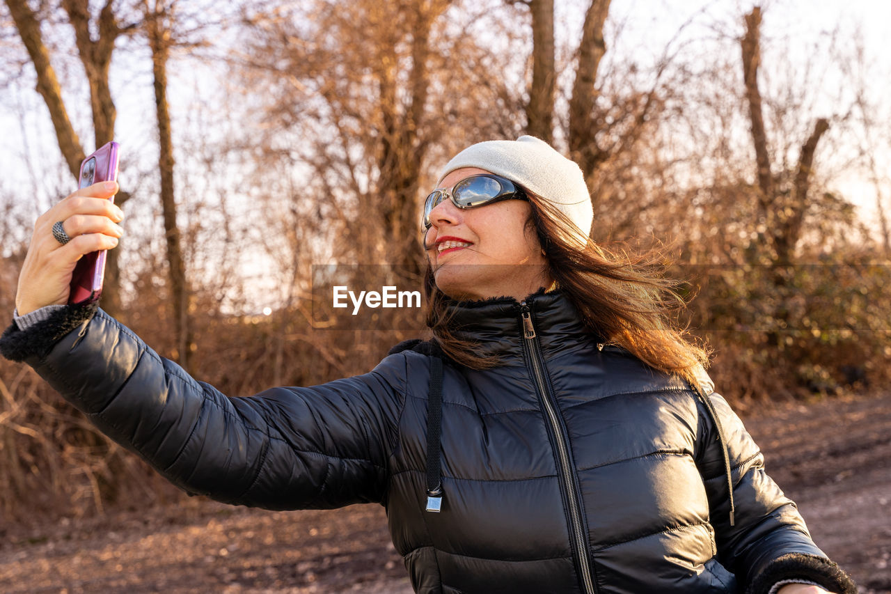 Middle aged woman wearing winter clothes taking selfie by a river - concept of people in recreation