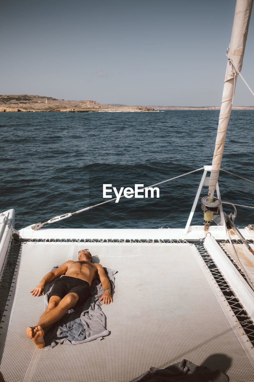 Full length of shirtless man lying on catamaran against sky during sunny day