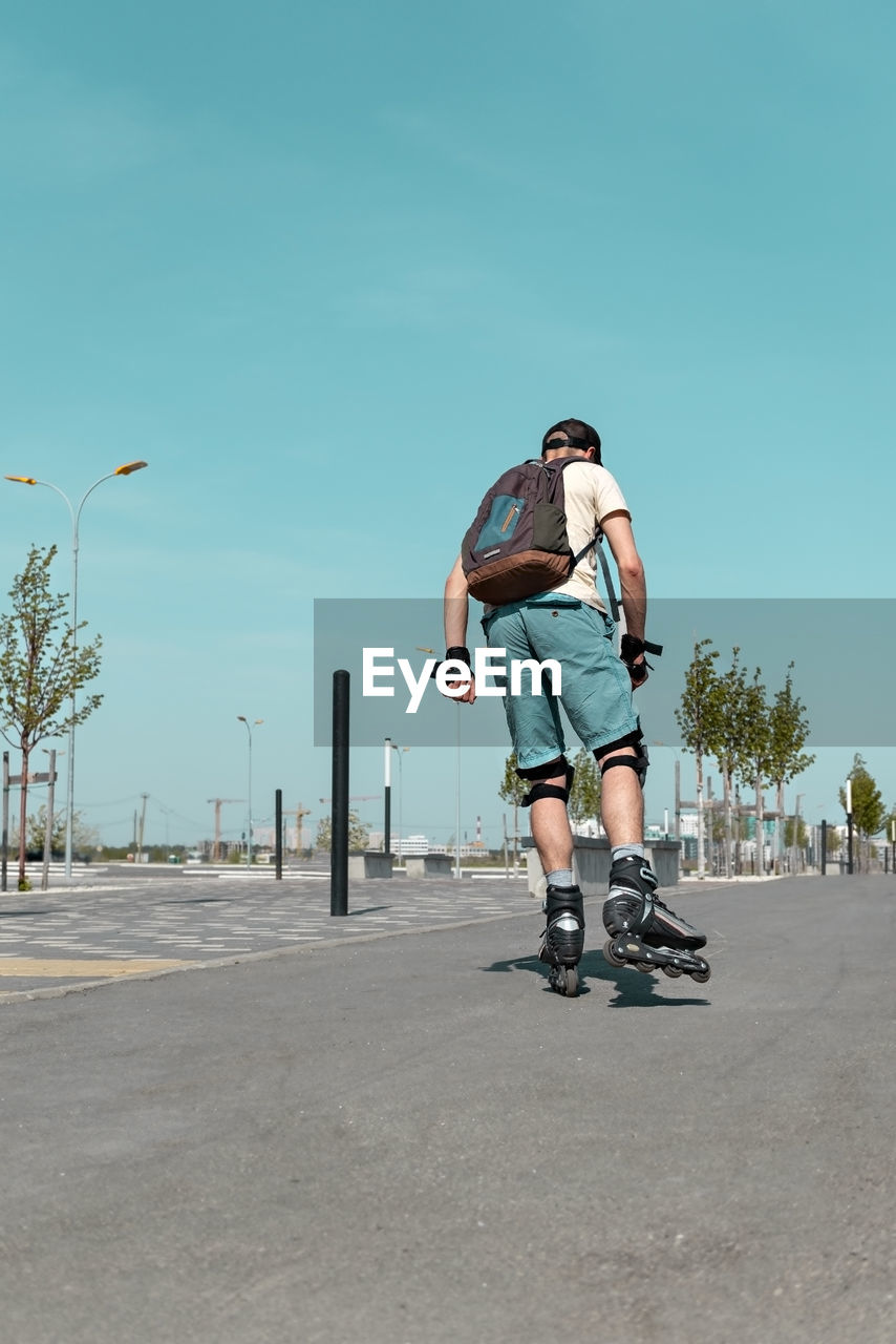 Rear view of young man with backpack in riding on roller skates on road in summer, roller blading