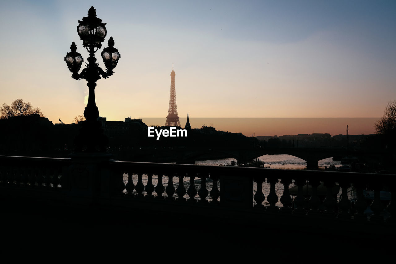 Silhouette lamp post on bridge with eiffel tower in background against sky during sunset