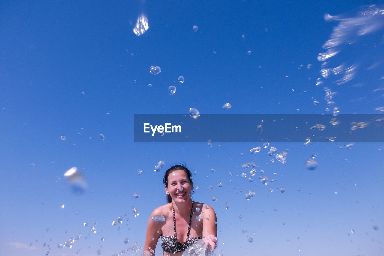 Smiling woman splashing water against clear blue sky during sunny day