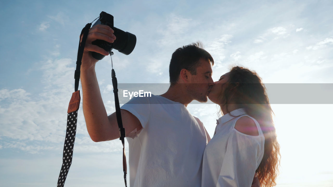 Young couple kissing on mouth against sky during sunny day