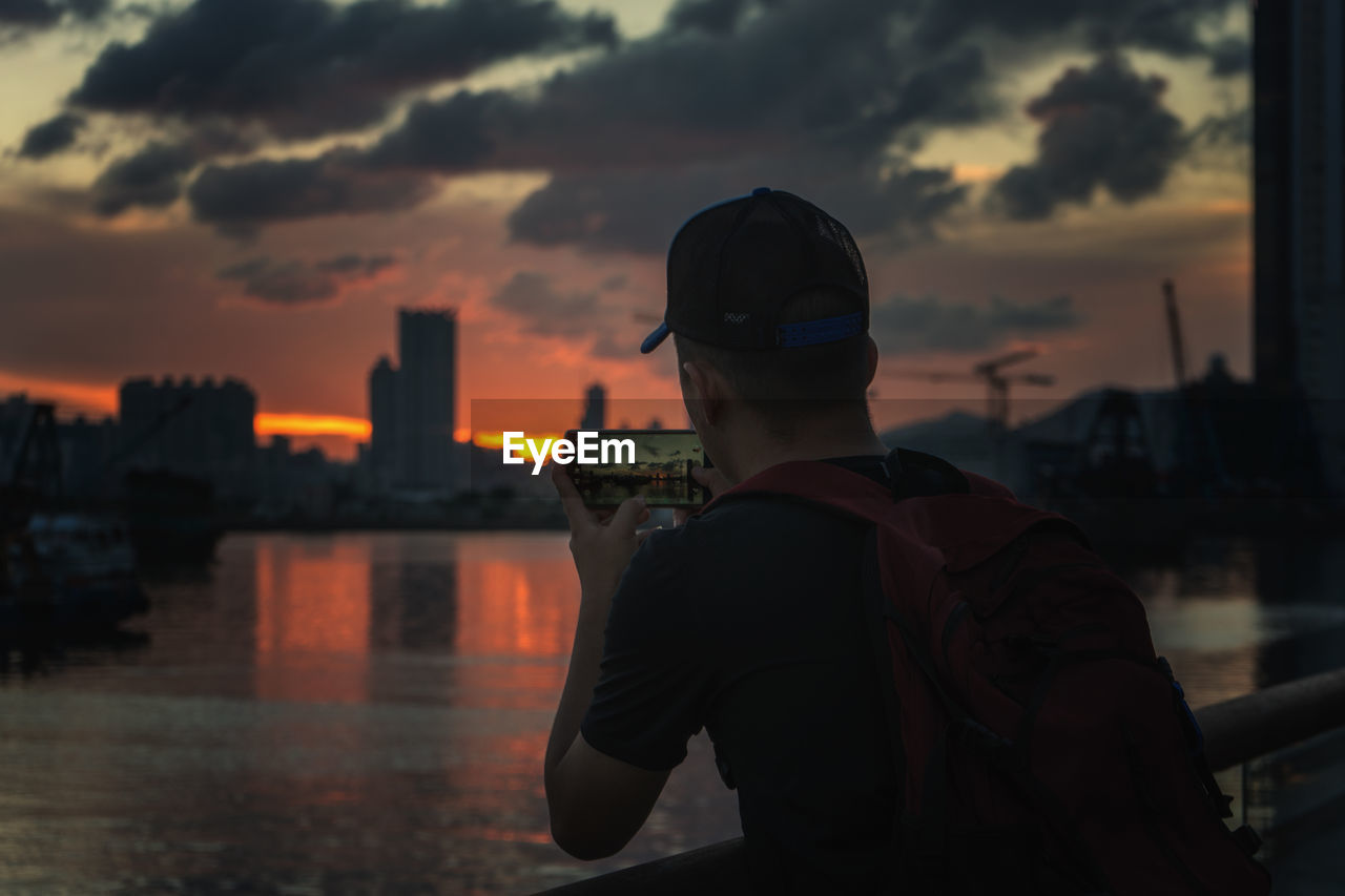 Rear view of man photographing sunset over city at kowloon bay