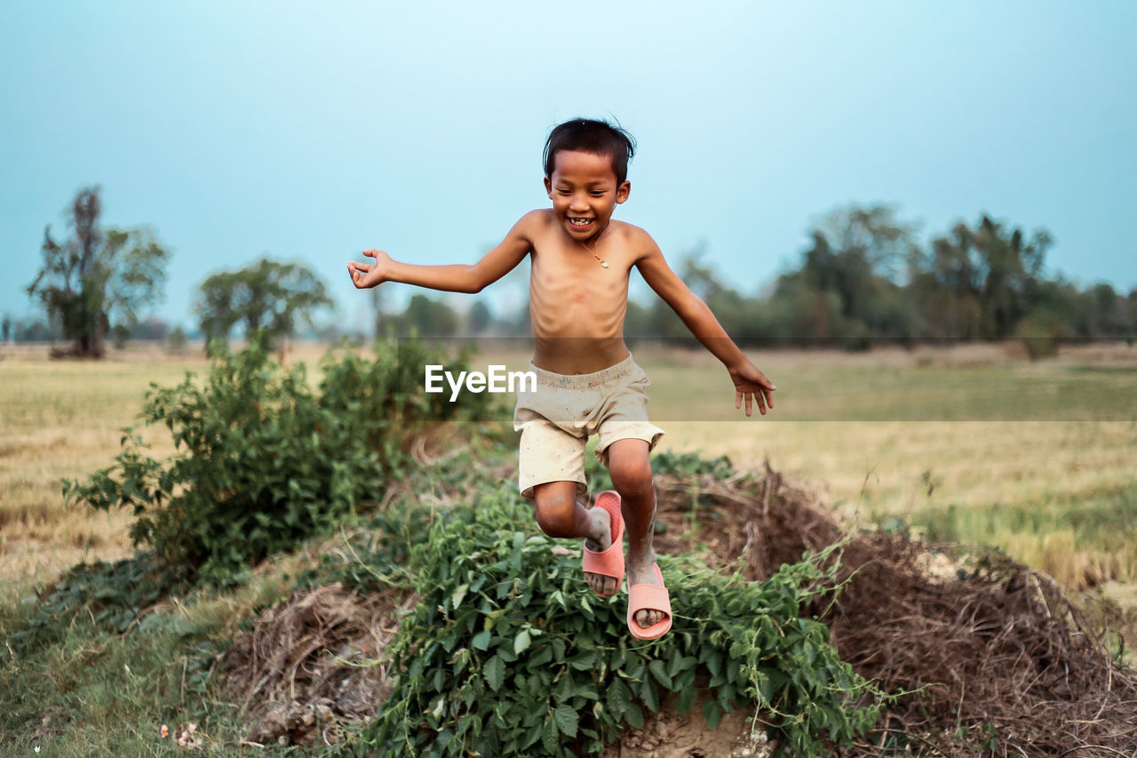 Happy shirtless boy jumping on field against sky at farm