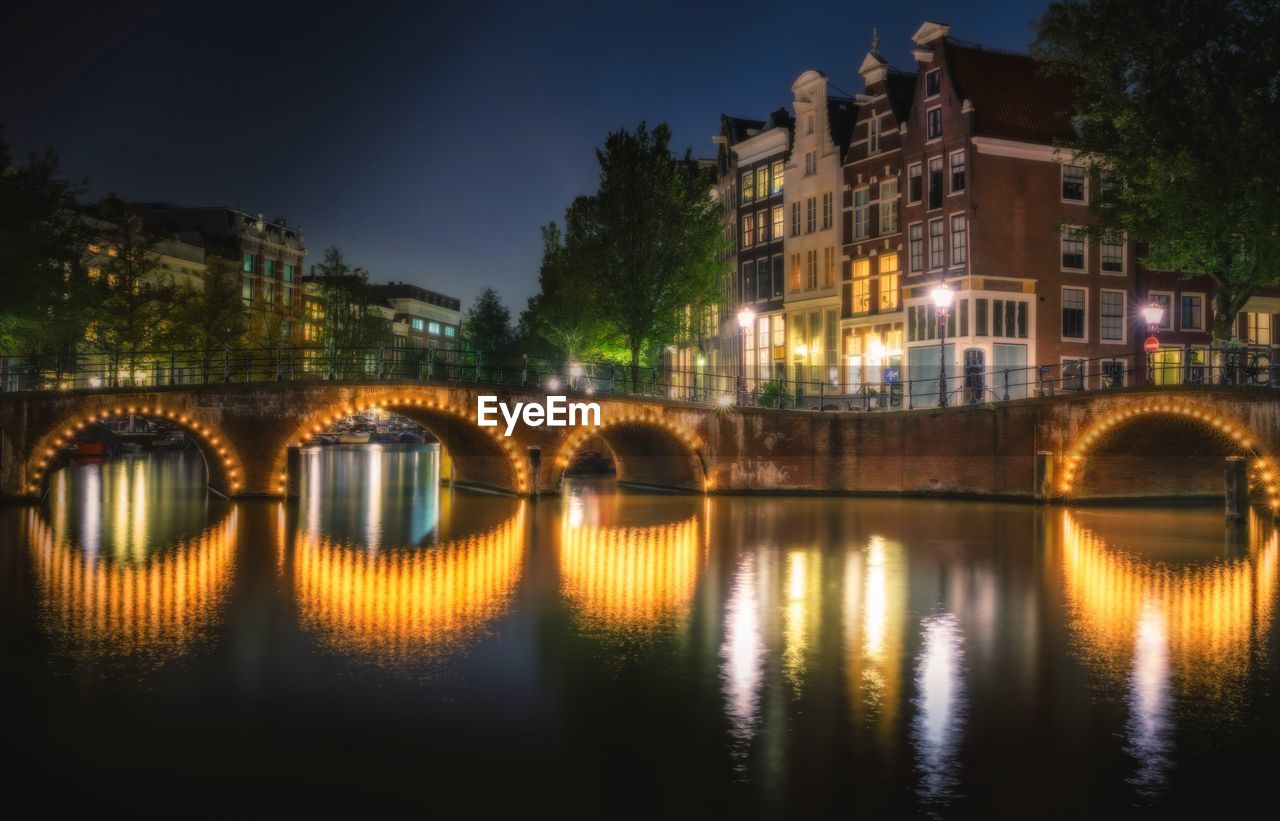 Illuminated bridge over river by buildings in city at night