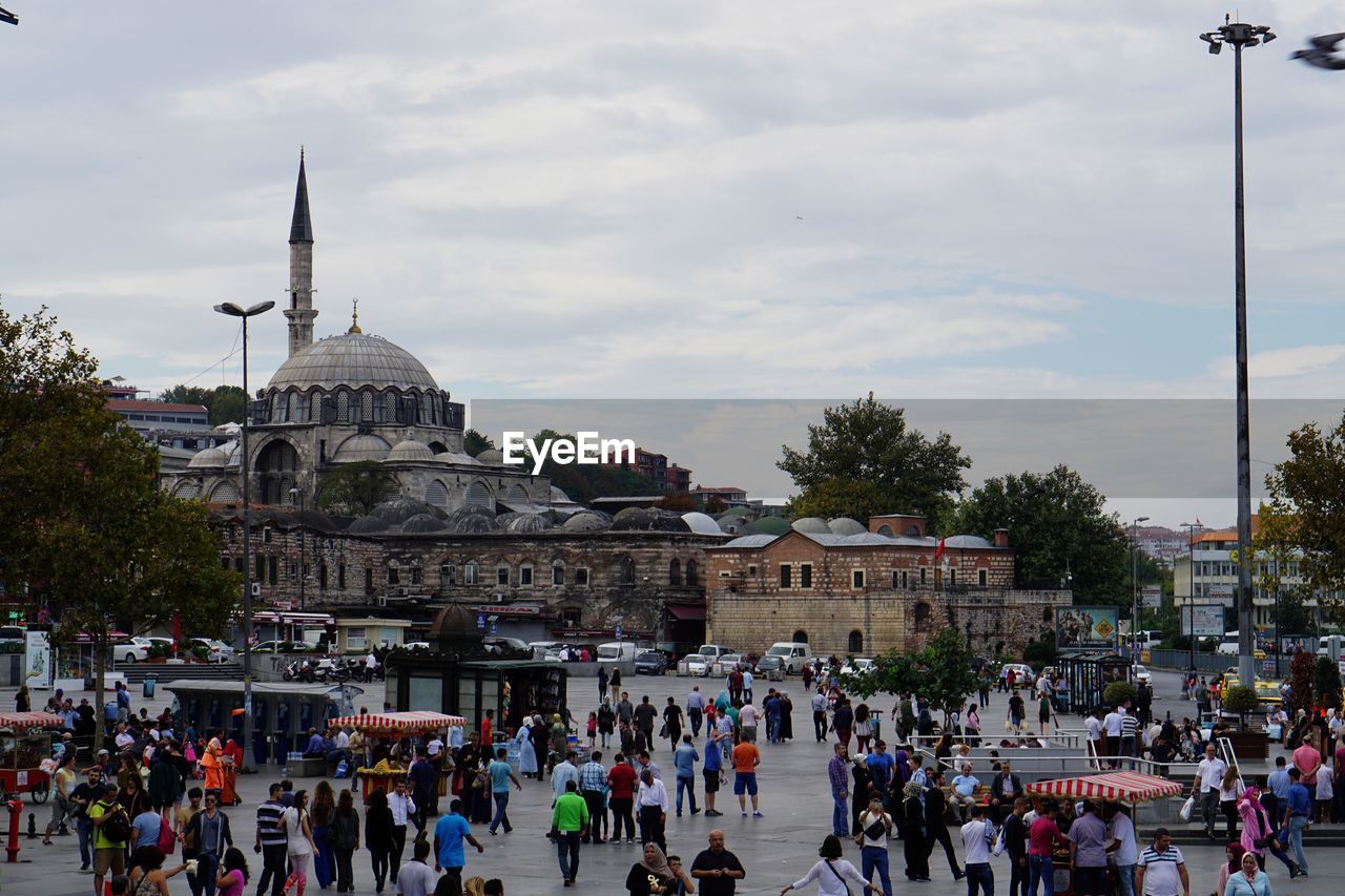 People walking on street by mosque against cloudy sky