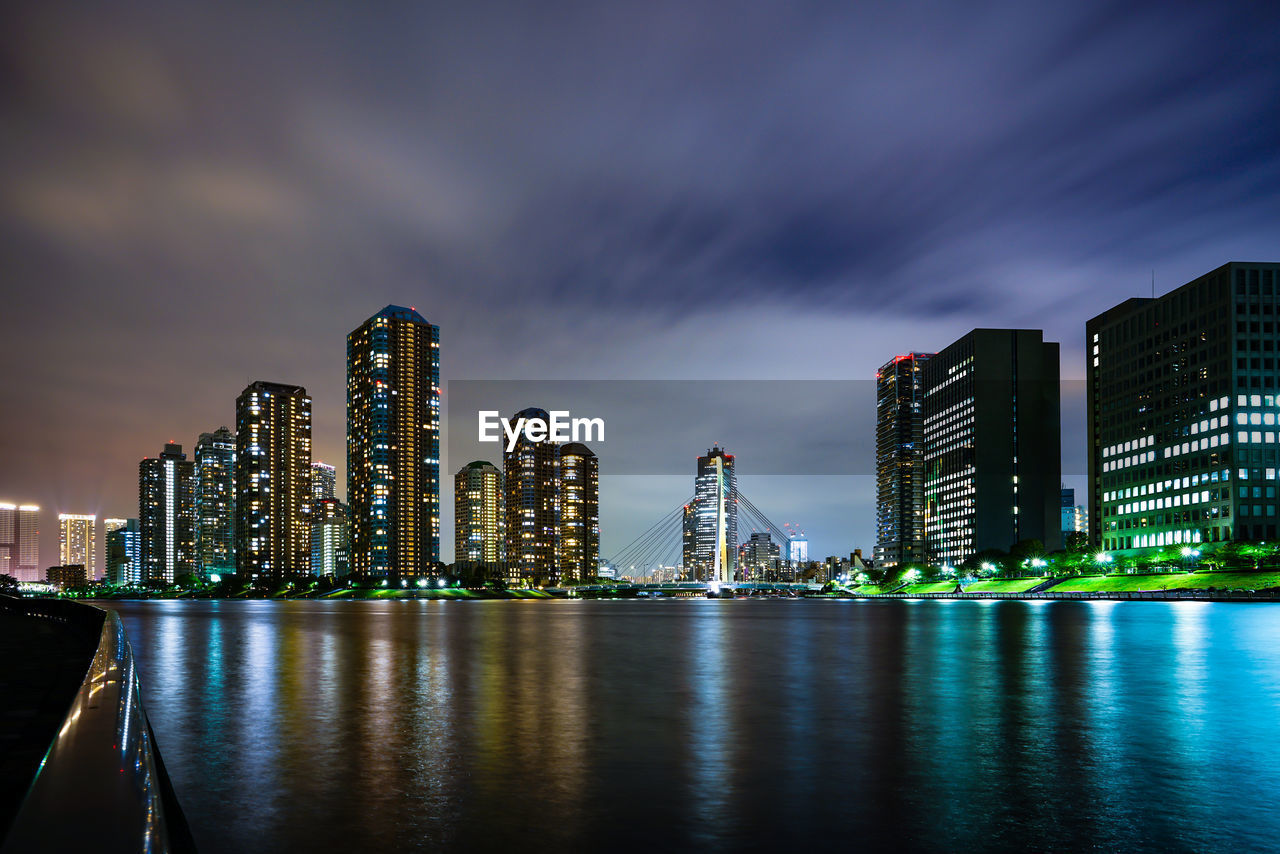 Illuminated modern buildings by bay against sky at night