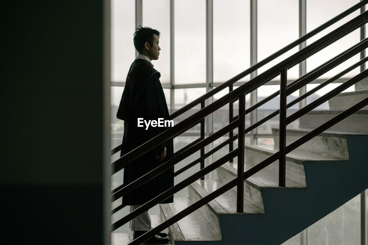 Full length serious man in graduation gown standing on stairs in building