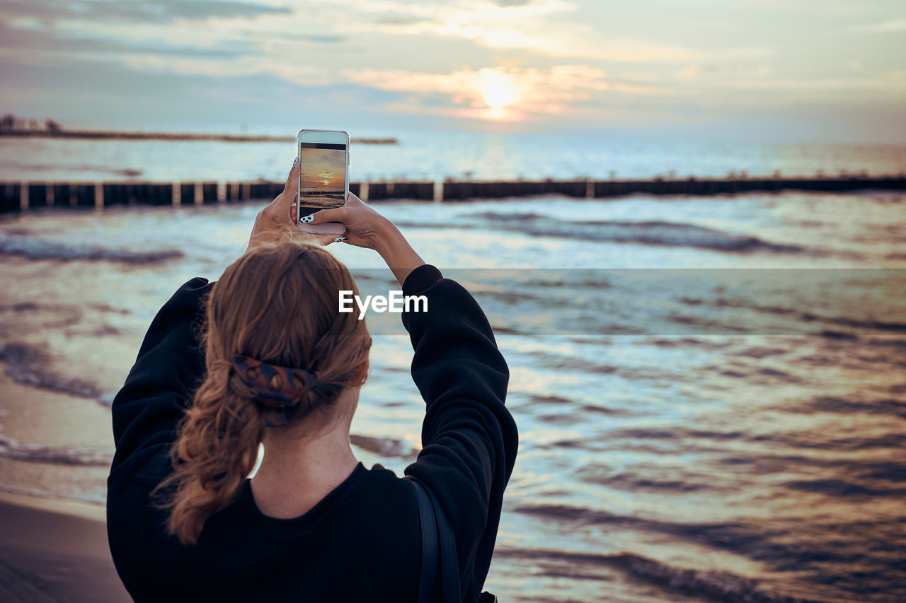 Young woman taking photos of sunset over sea using smartphone during summer trip