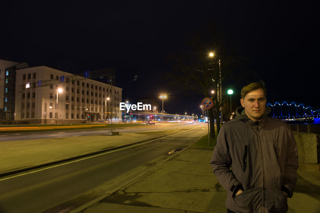 Portrait of young man standing on sidewalk in city at night