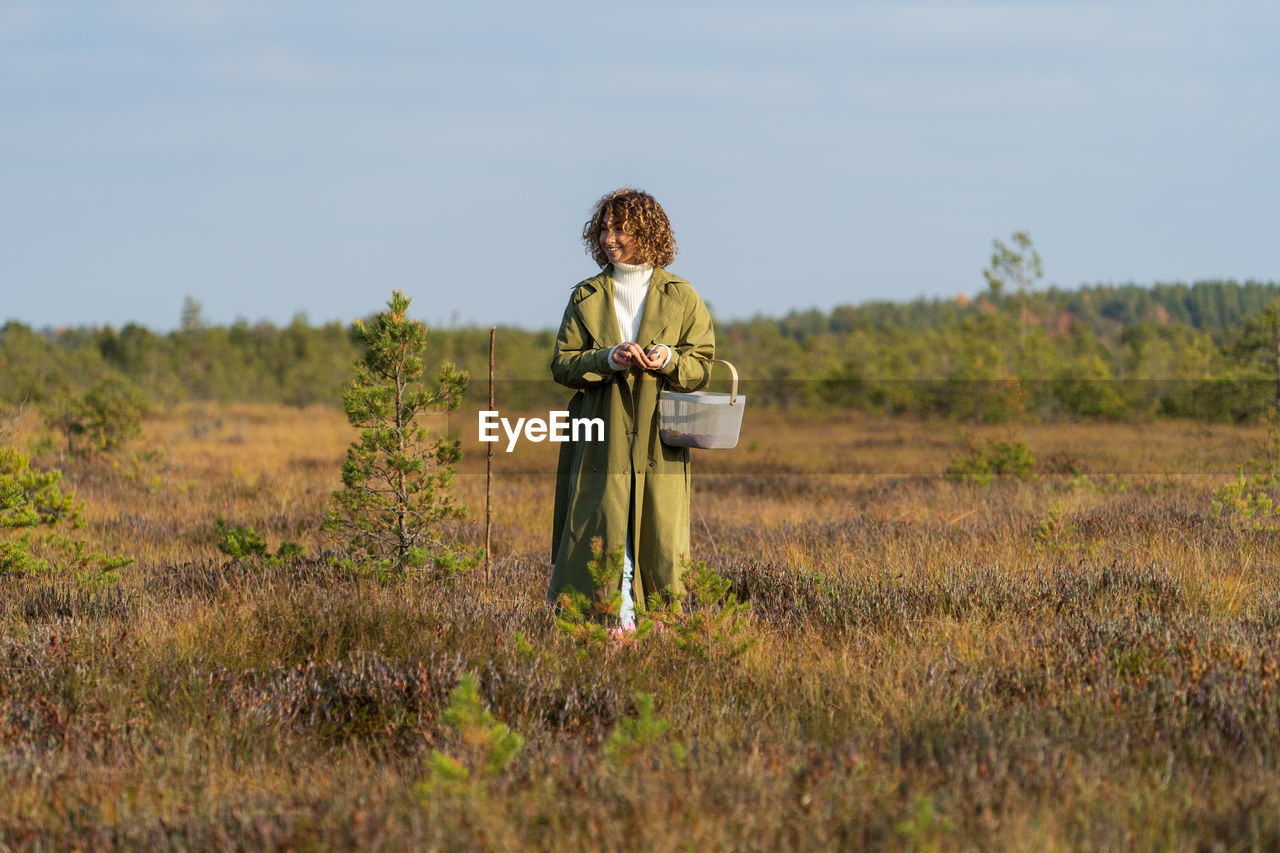 Happy lady in trench coat looks for cranberries among grass resting in autumn nature