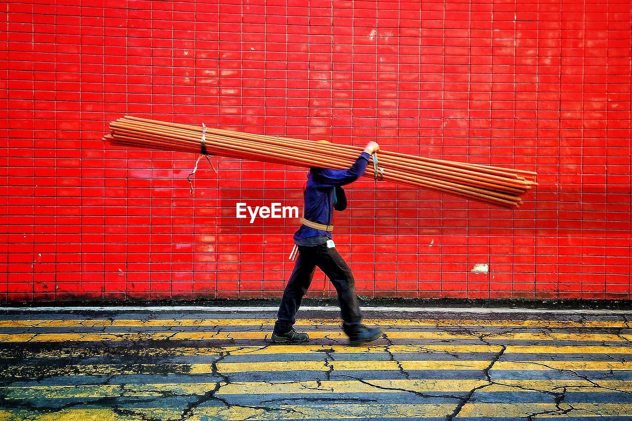 Side view of man carrying metallic rods while walking against wall