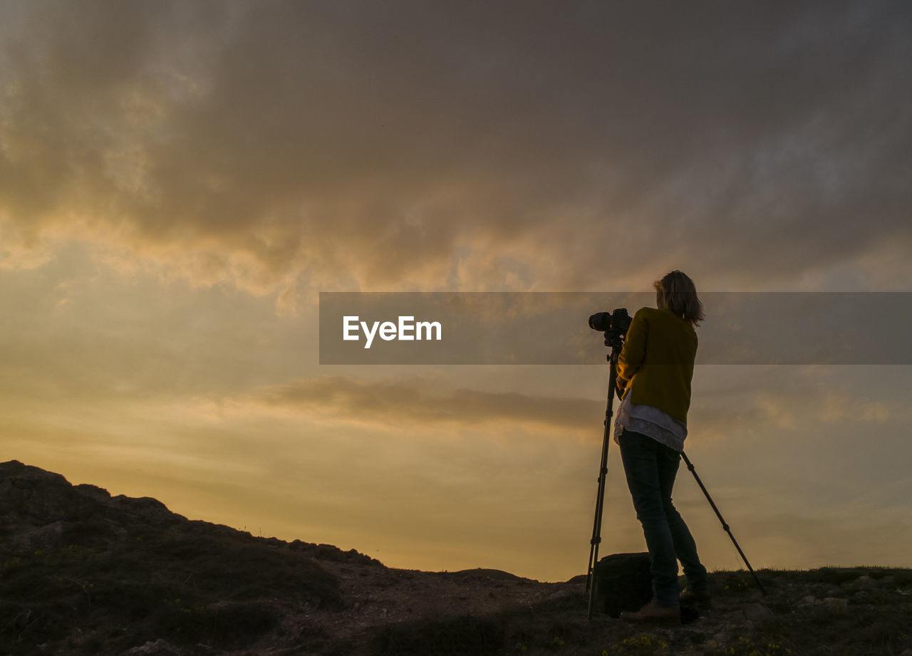 Low angle view of female photographer standing by tripod against cloudy sky during sunset