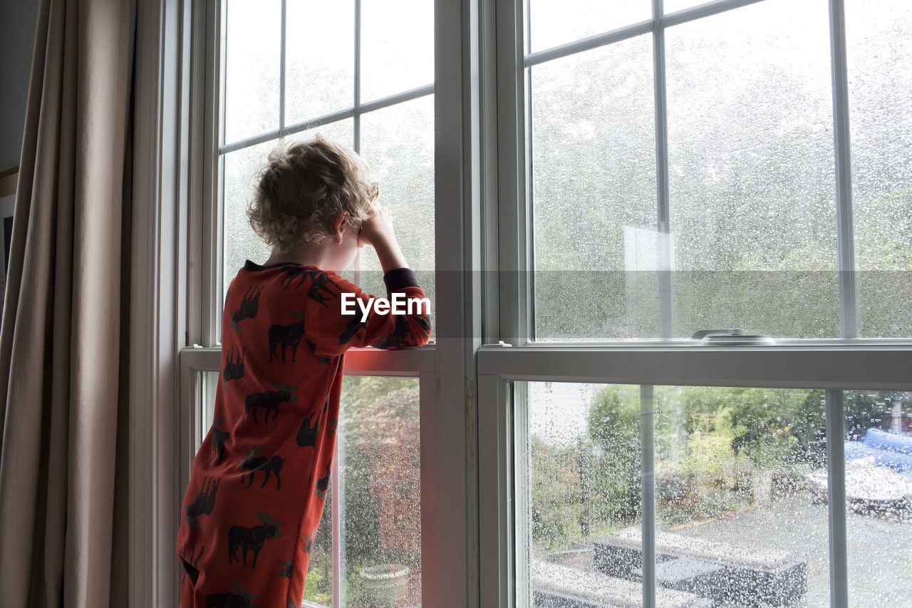 Rear view of young boy with curly hair looking out rain covered window