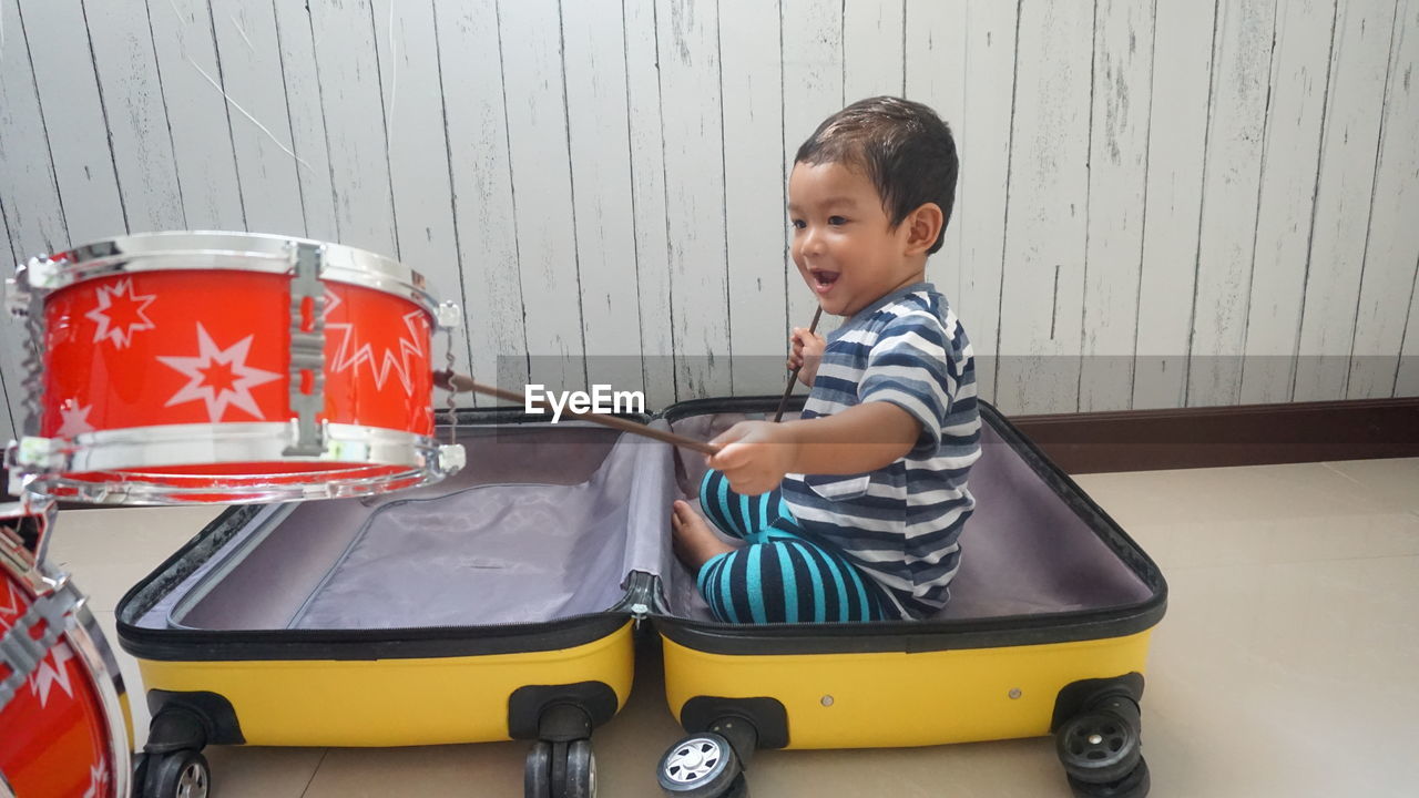 Cute boy sitting in luggage by drums at home