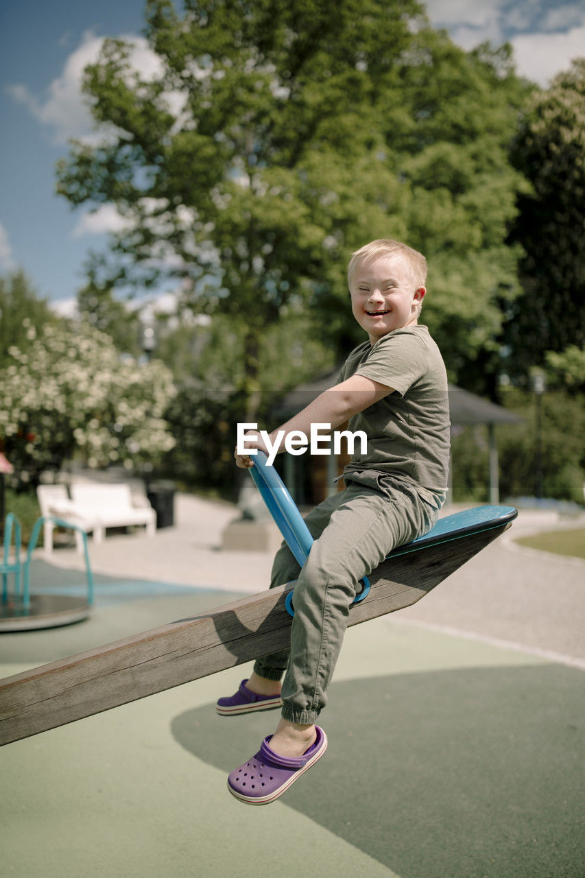 Full length side view portrait of happy boy with down syndrome sitting on seesaw at park