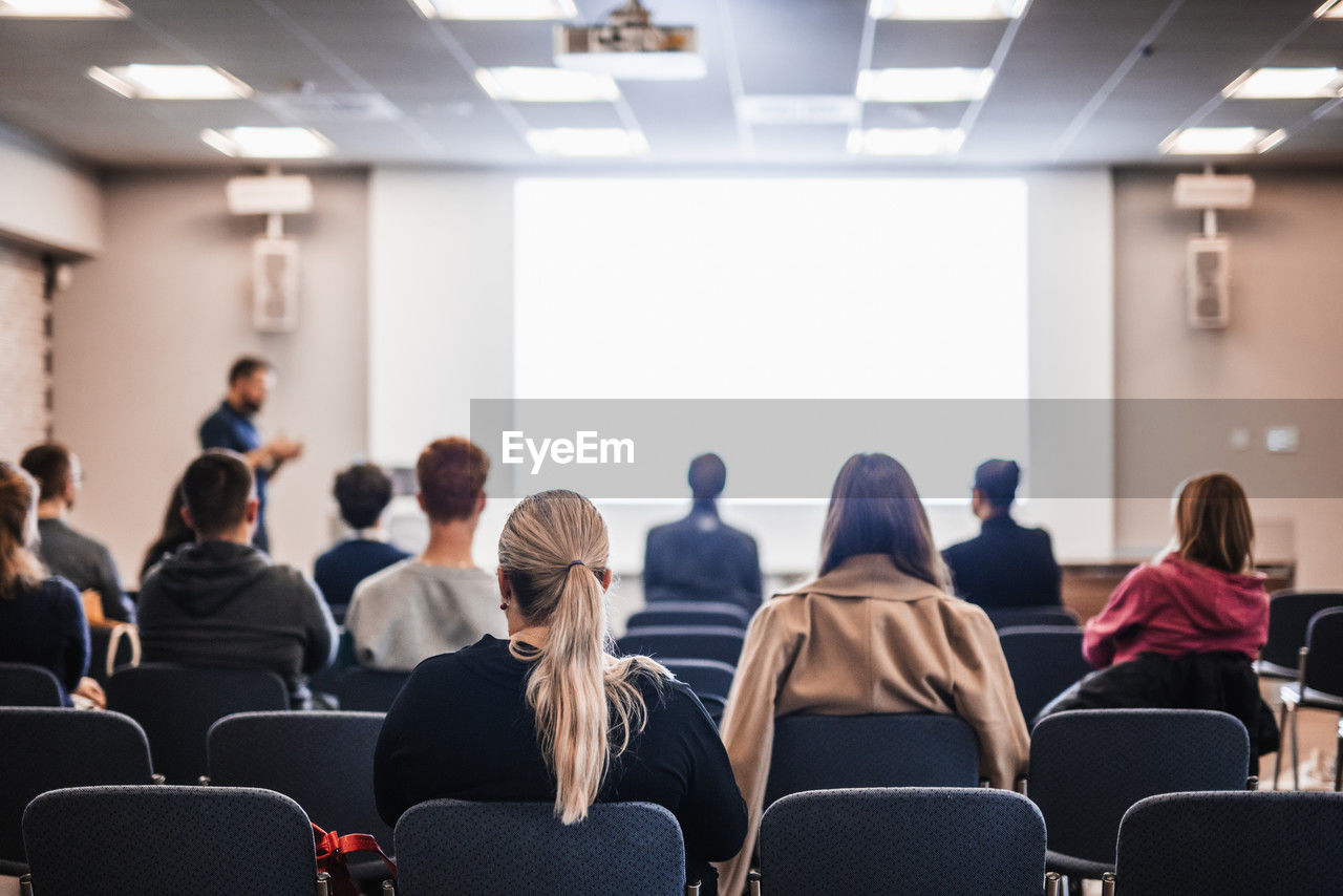 group of people, adult, women, indoors, meeting, sitting, men, seminar, female, crowd, auditorium, communication, lecture hall, business, large group of people, business meeting, talking, audience, student, learning, convention center, education, university student, seat, university, young adult, mature adult, business conference, businesswoman, businessman, corporate business, cooperation, copy space, listening, conference hall, rear view, discussion, chair, teamwork, adult student, convention, school, looking, office, conference, watching, togetherness