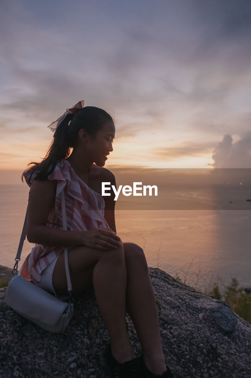 Side view of young woman sitting on beach against sky during sunset