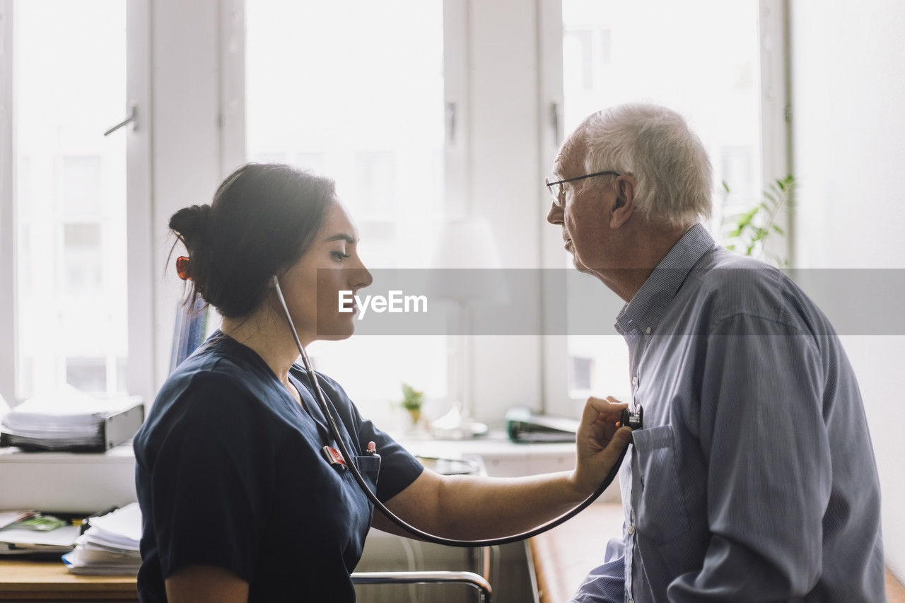 Side view of female nurse listening heartbeat of senior male patient in clinic