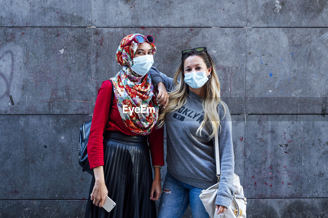 Woman wearing face mask while standing with hand on muslim friend's shoulder against gray wall during pandemic