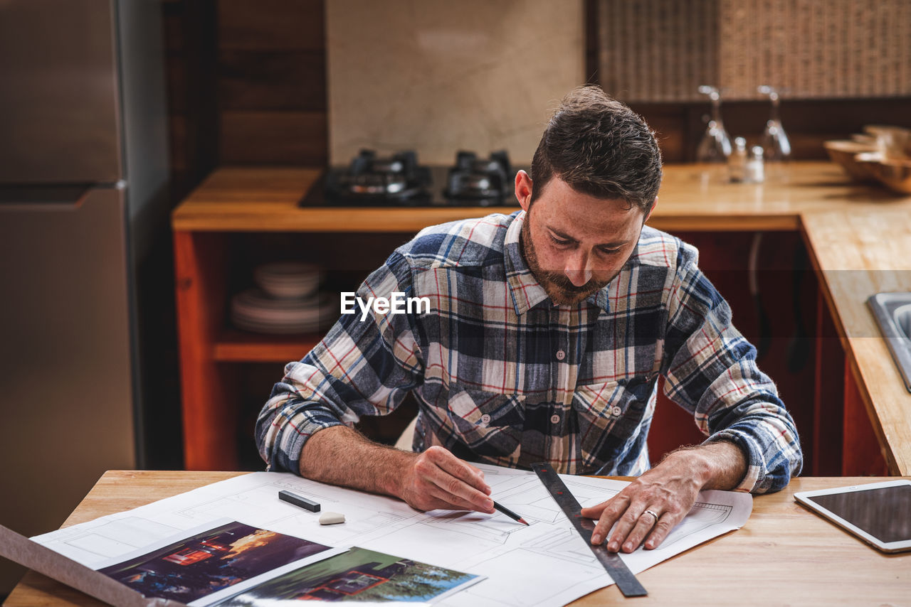 Serious male architect sitting at table in kitchen and drawing blueprint of building with pencil and ruler while working at home