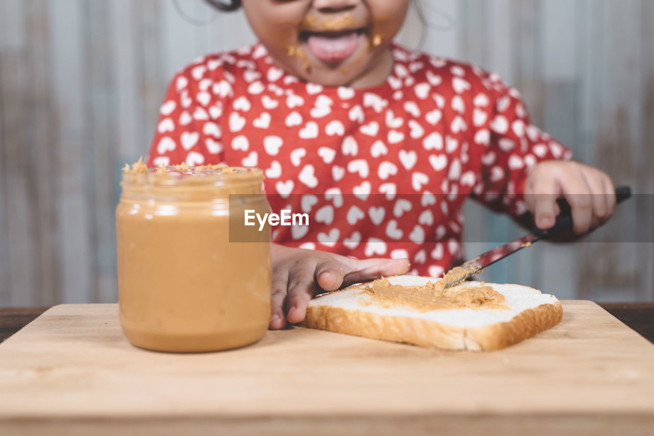 Midsection of girl applying peanut butter on bread at table against wall