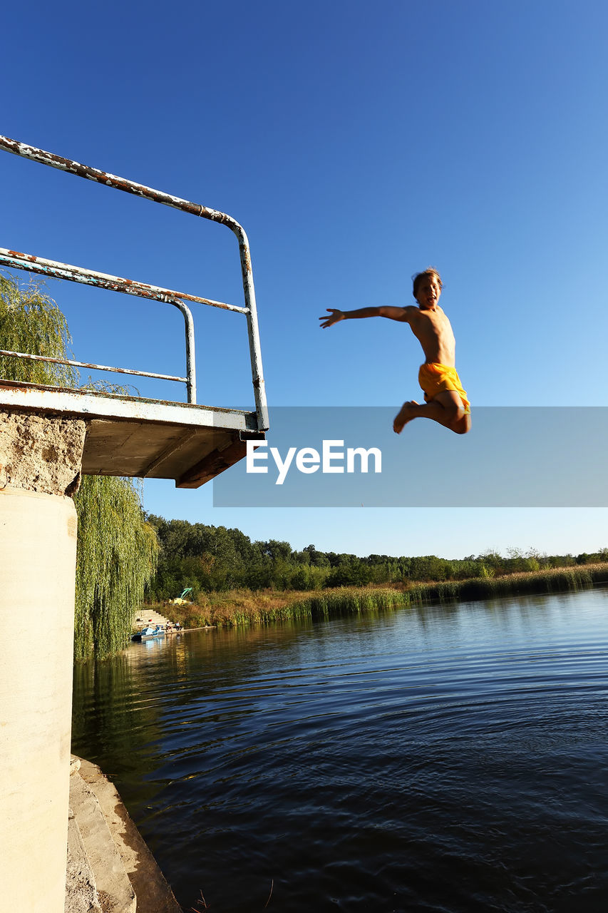 LOW ANGLE VIEW OF MAN JUMPING OVER LAKE AGAINST CLEAR SKY