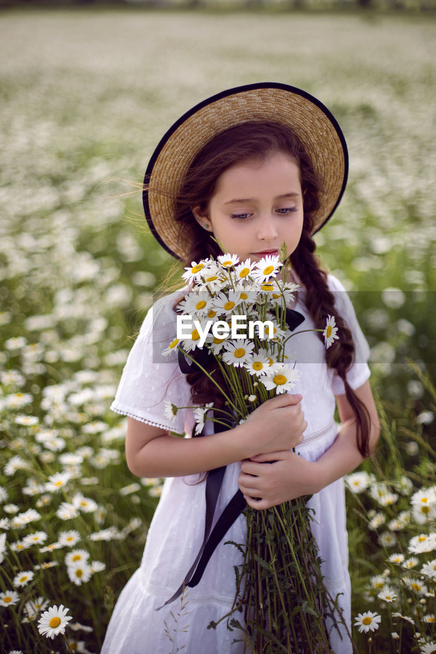 Portrait girl child in a white dress stands on a camomile field in a hat. bouquet of flowers