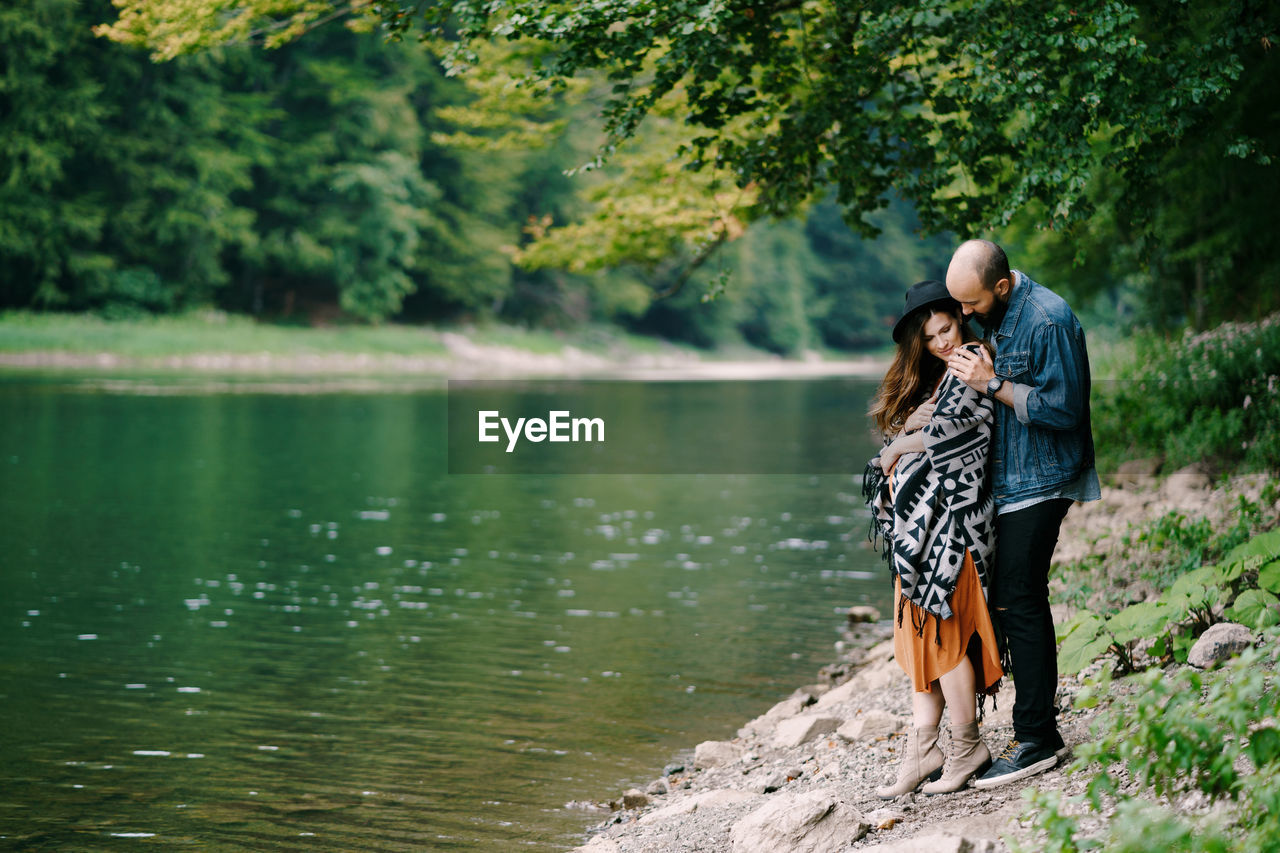Couple standing by lake against trees