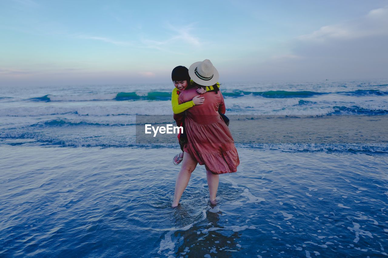Mother carrying son while standing at beach against sky