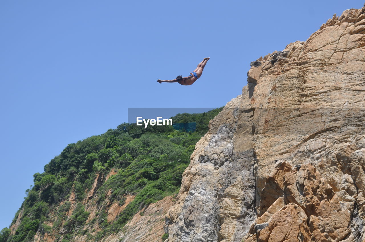 LOW ANGLE VIEW OF MAN JUMPING FROM ROCK AGAINST CLEAR BLUE SKY