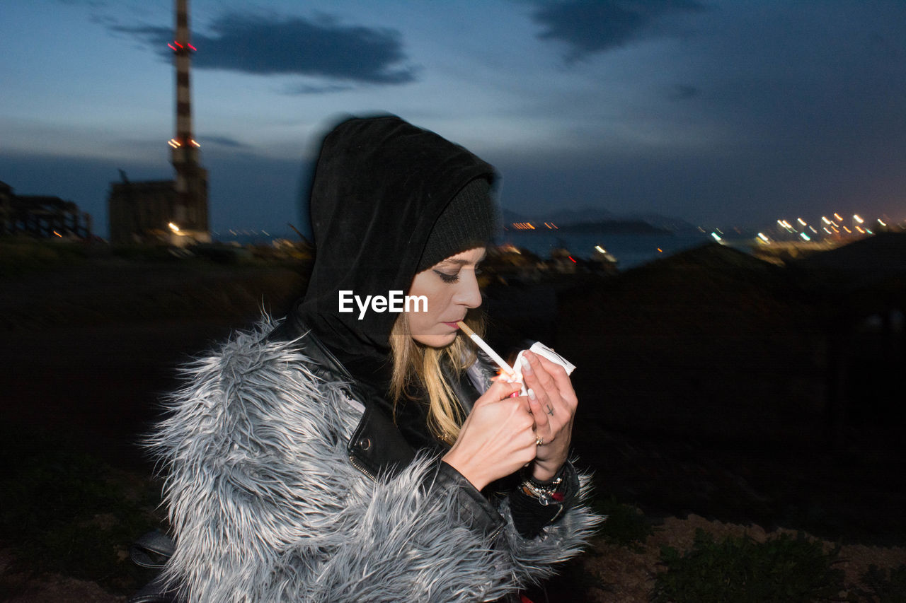 Young woman lighting cigarette in city against sky at night