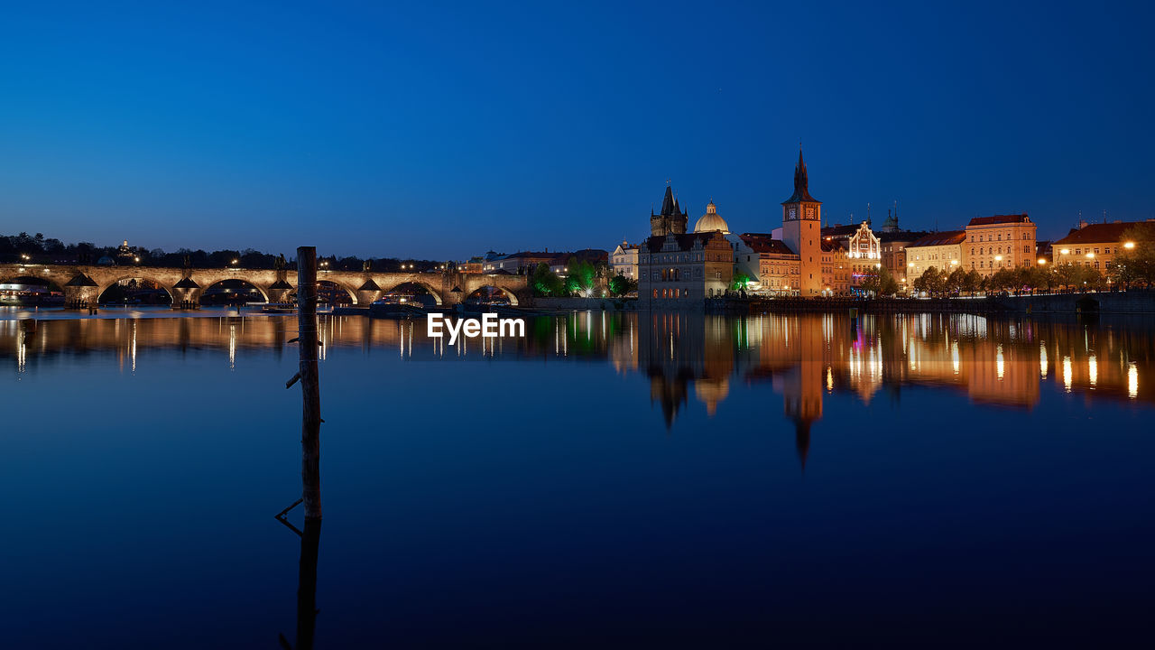 Bridge over river by illuminated town against clear blue sky