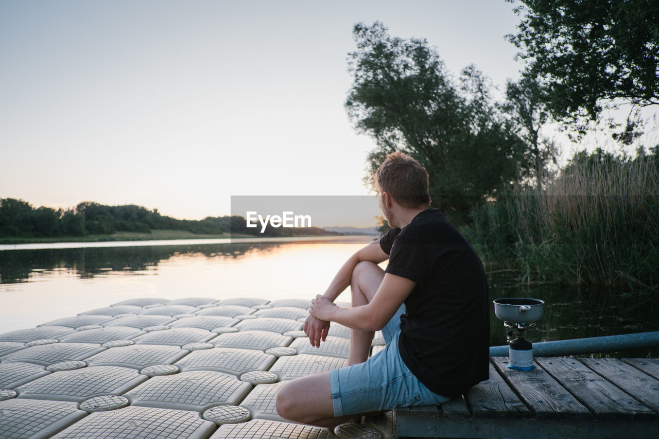 MAN SITTING BY LAKE AGAINST SKY