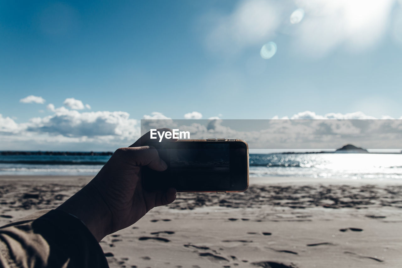 Hand holding smart phone at beach against sky