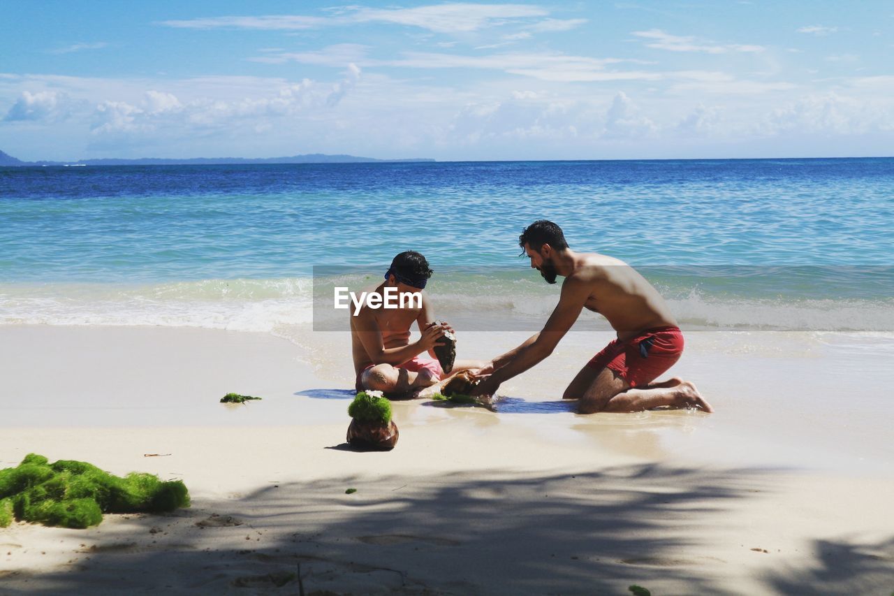 Shirtless men breaking seashell at beach against sky