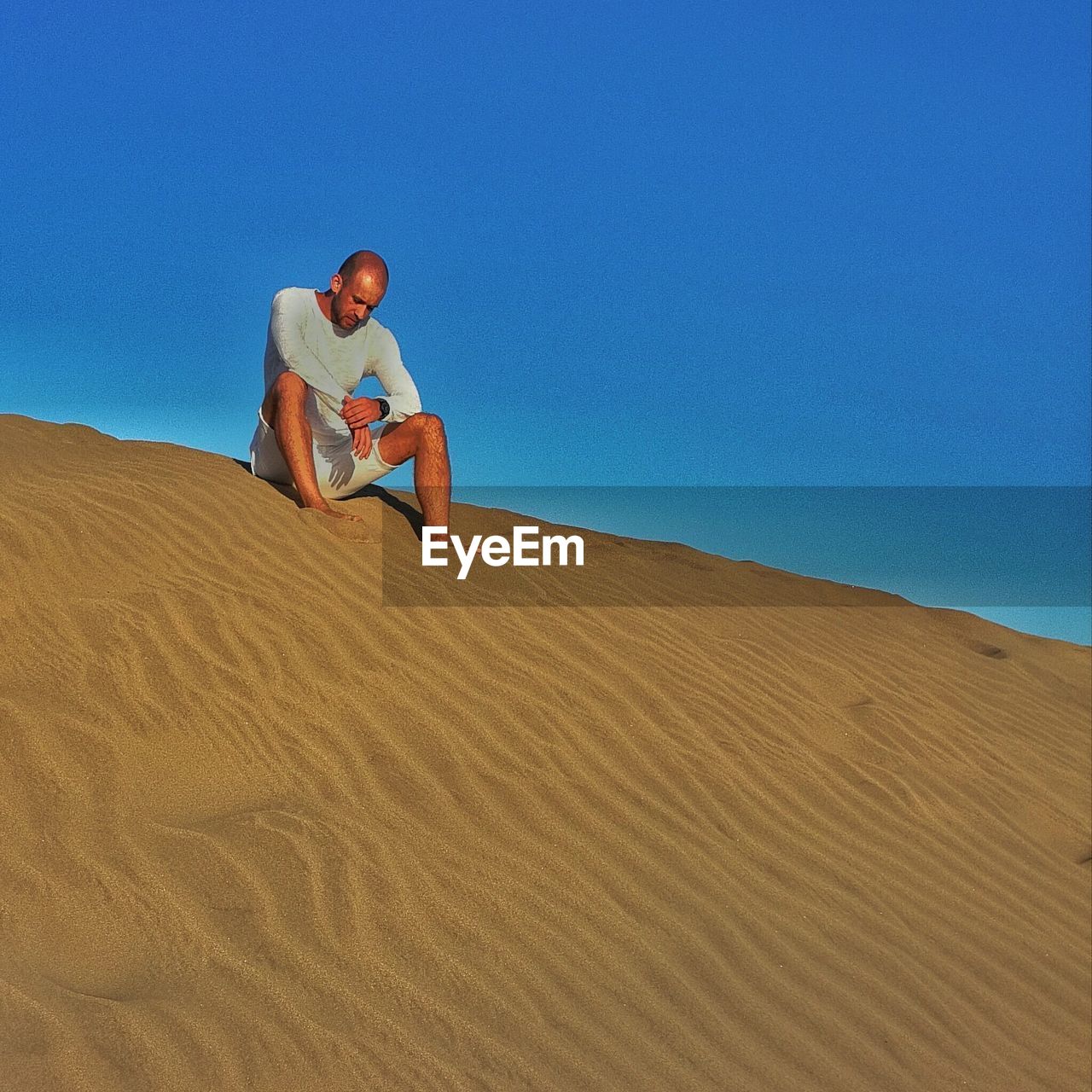 Low angle view of tired man sitting on sand dune against clear blue sky