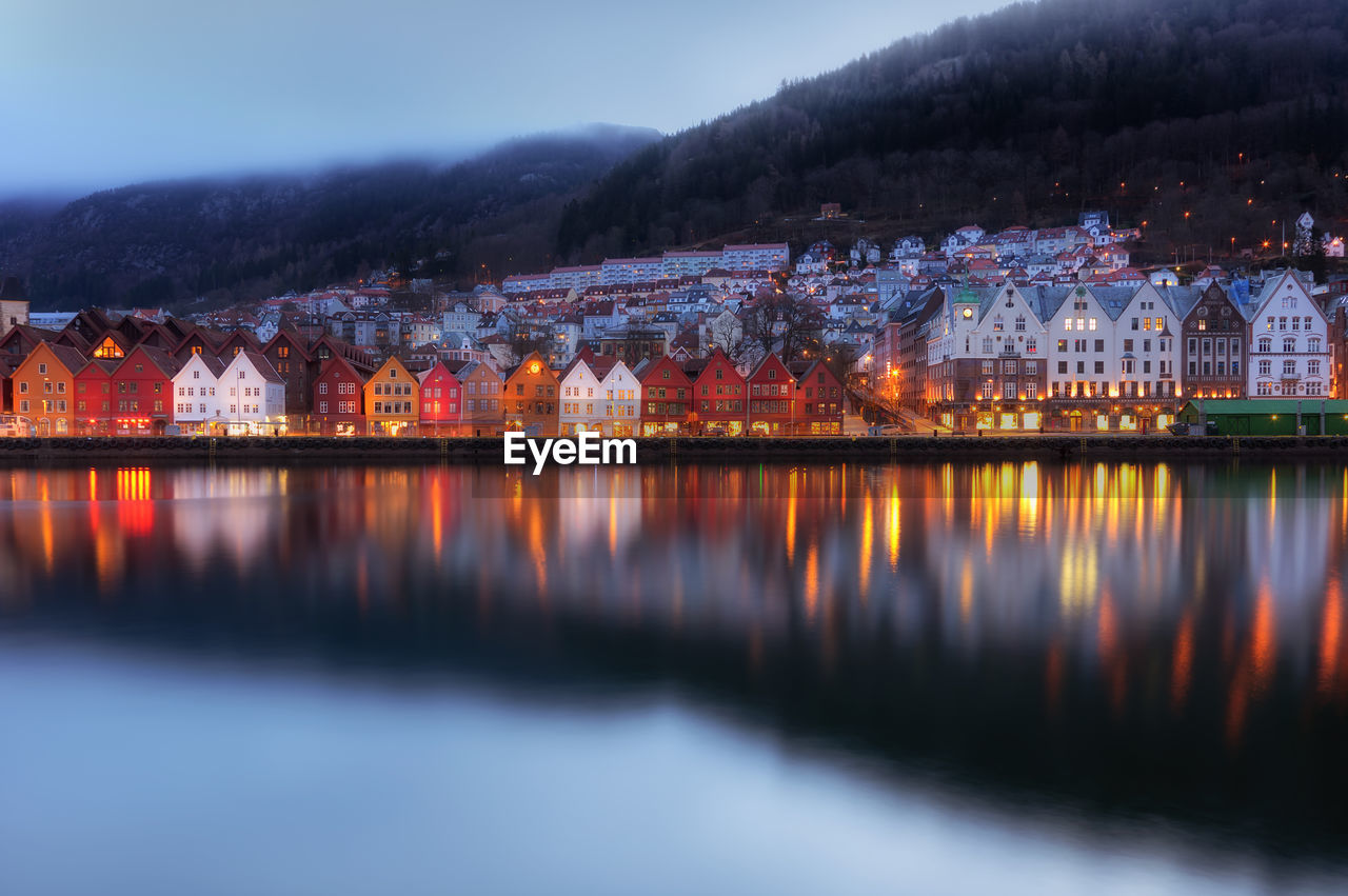 Illuminated buildings by lake against sky at dusk