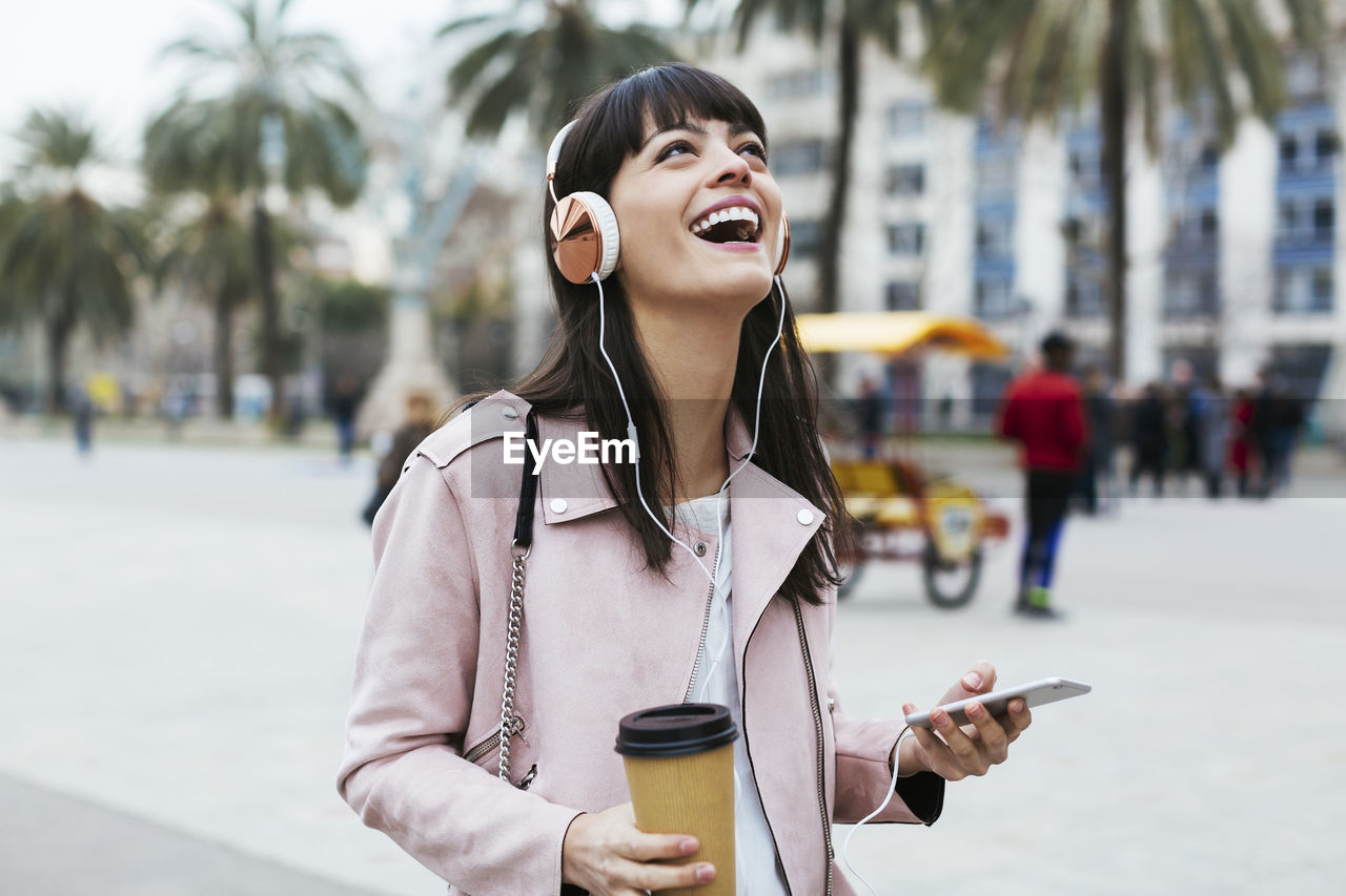 Spain, barcelona, laughing woman with coffee, cell phone and headphones in the city