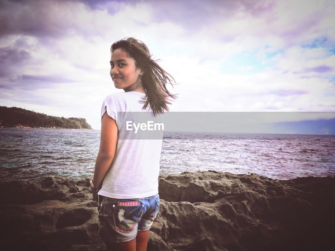 Portrait of young woman standing on rocky shore at beach against cloudy sky