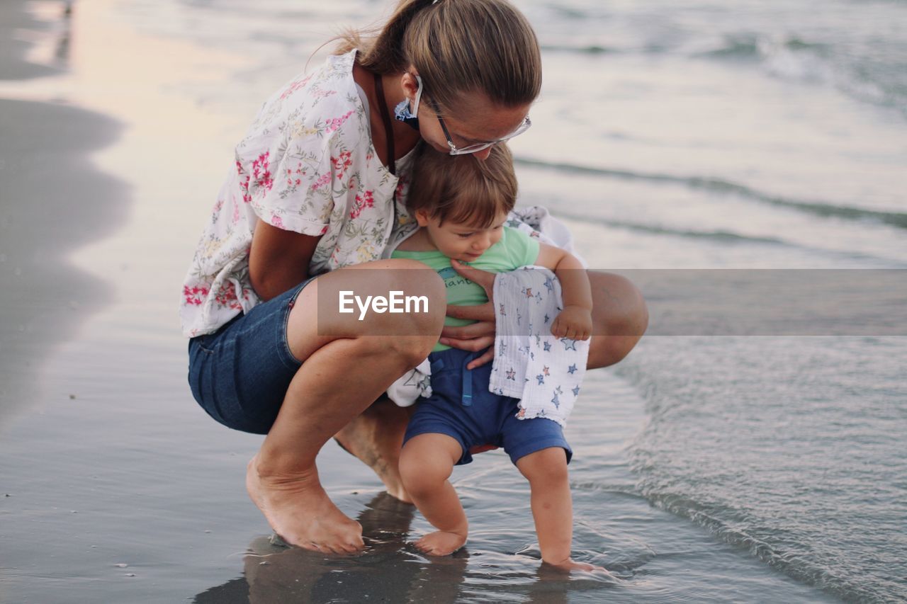 Rear view of mother and cute blonde baby boy on the beach touching the sea for the first time