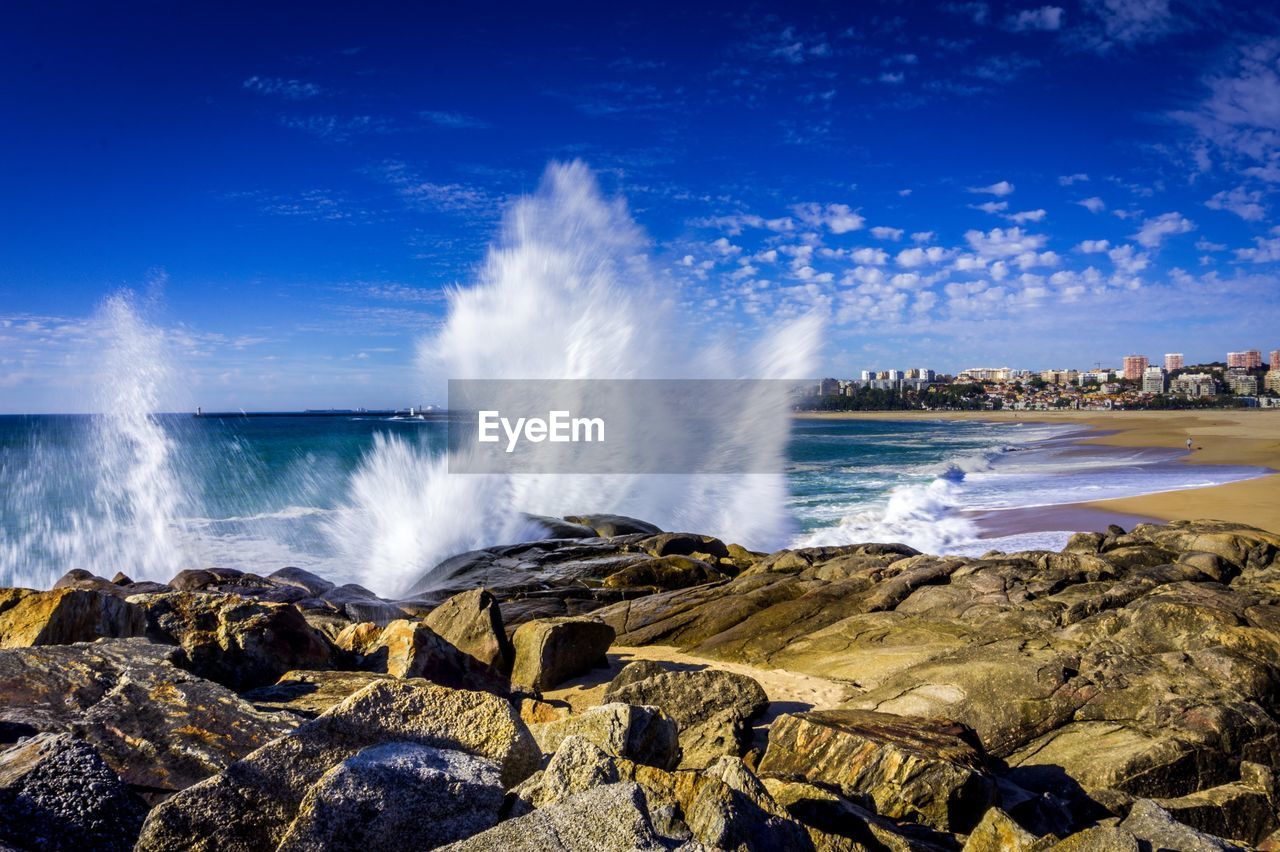 Waves splashing on rocks at shore against blue sky