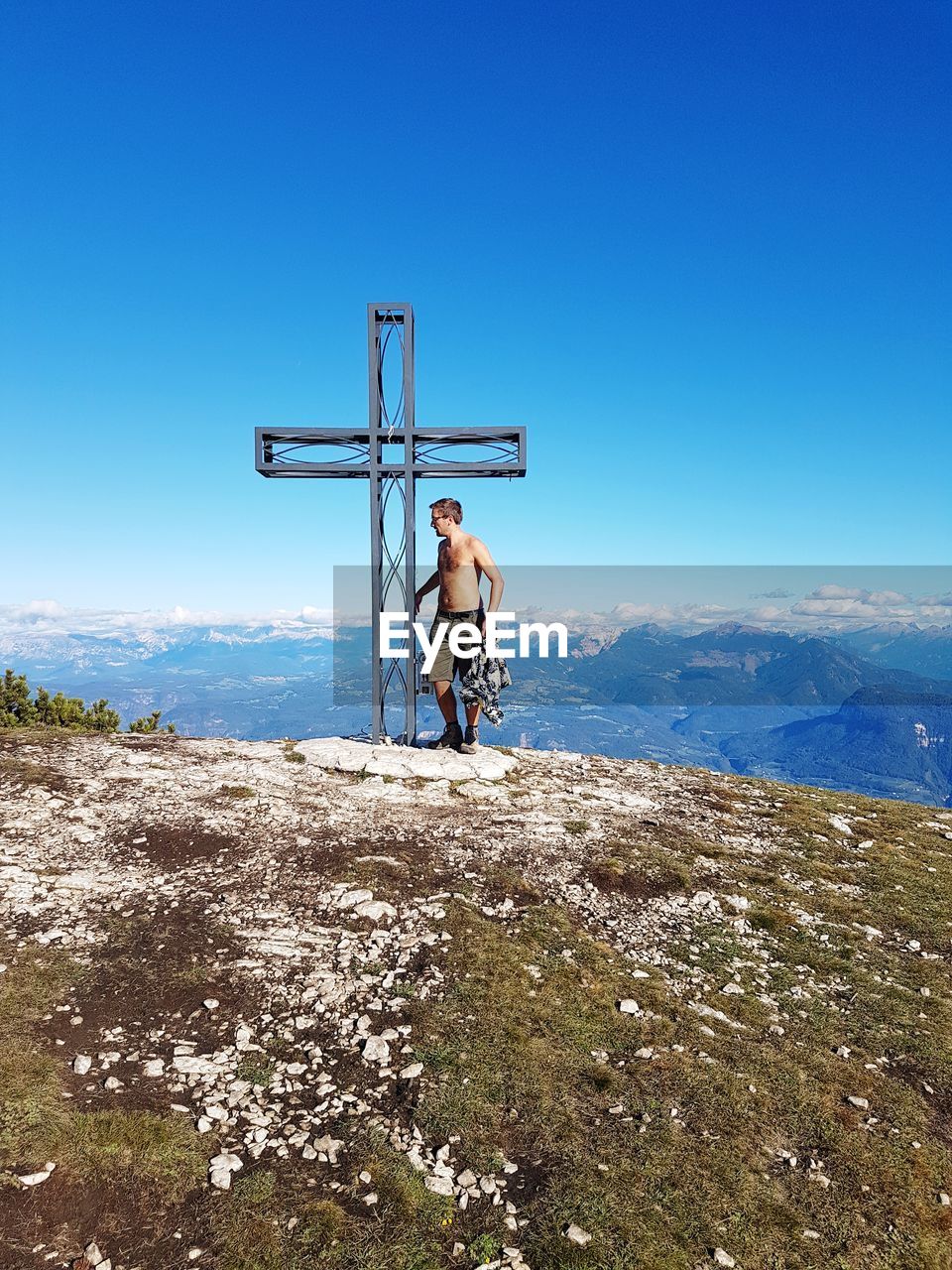 Shirtless man standing by cross on mountain against clear blue sky