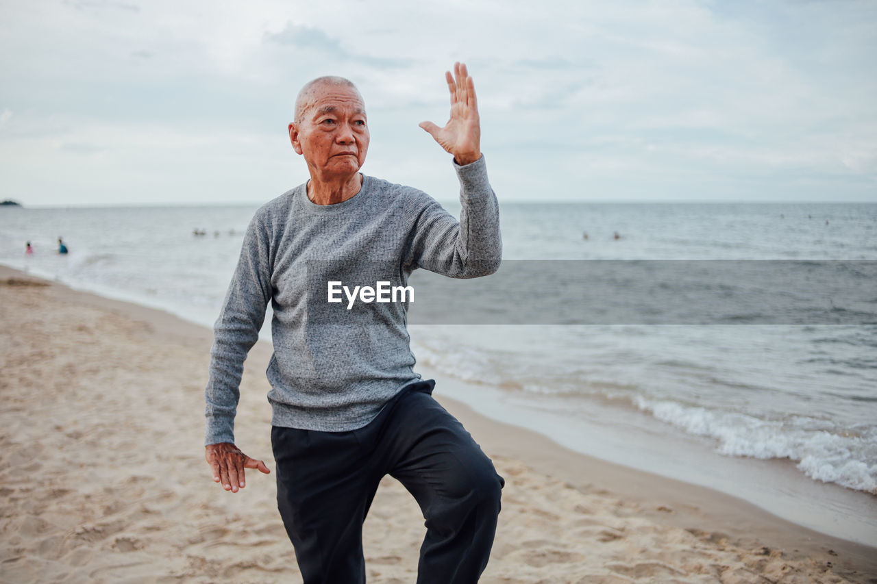 Senior man exercising while standing at beach against cloudy sky during sunset