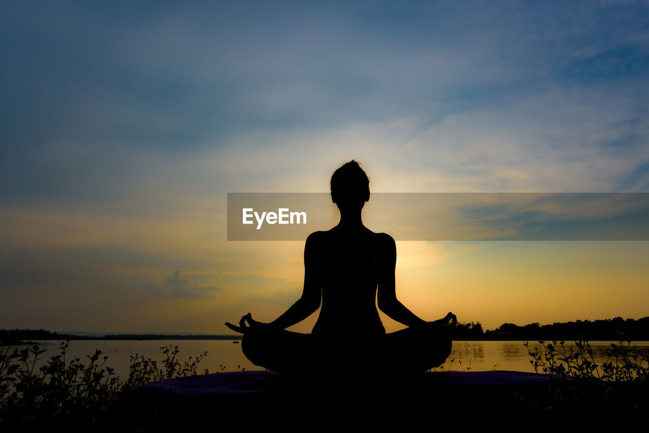 Silhouette woman meditating in lotus position at lakeshore against sky