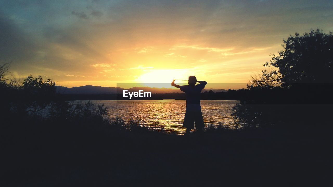 Silhouette boy standing by lake against sky during sunset