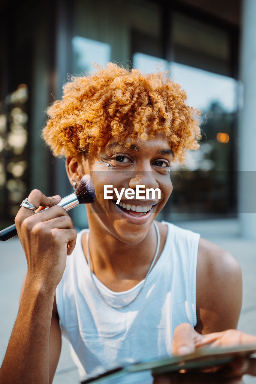 Positive multiracial charismatic boy preparing makeup at his face with visage brush